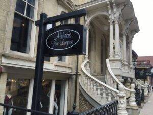 The image shows the exterior of Althea's Fine Lingerie, located at 777 N. Jefferson Street in Milwaukee. The building has a historic architectural style with light-colored brick walls and ornate columns. A grand, curved staircase with intricate railings leads up to the entrance. The black store sign with white lettering reads 'Althea's Fine Lingerie' and hangs from a post attached to the building. A glimpse of the storefront windows reveals part of the interior display, hinting at a cozy and elegant shopping experience. The overall scene is inviting and showcases the boutique's unique, upscale setting.